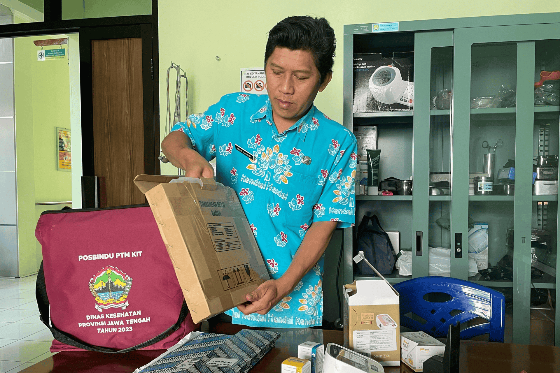 Person opening a  box beside a maroon POSBINDU PTM KIT bag and medical supplies.