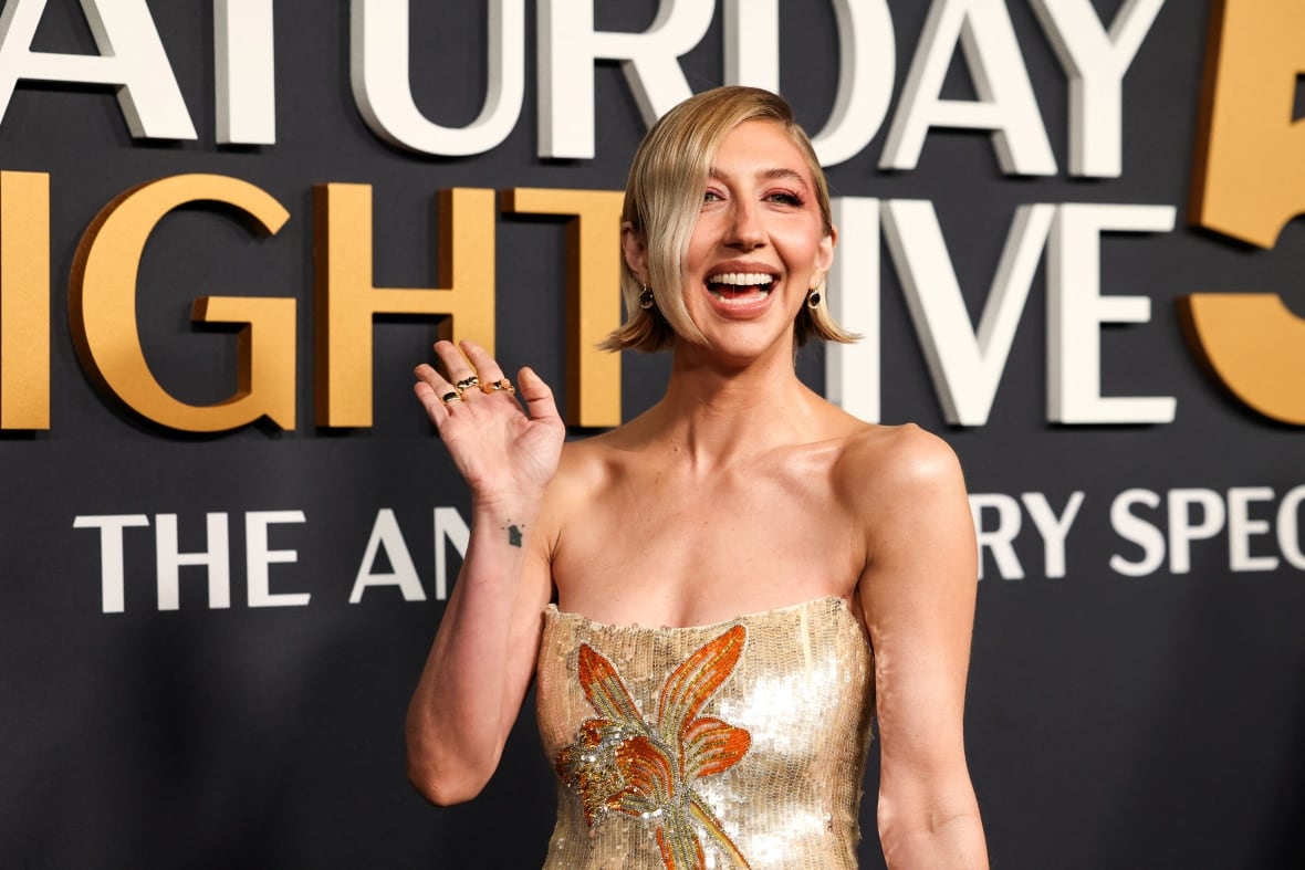 a woman with blonde hair in a gold strapless dress waves and smiles to the camera on a red carpet