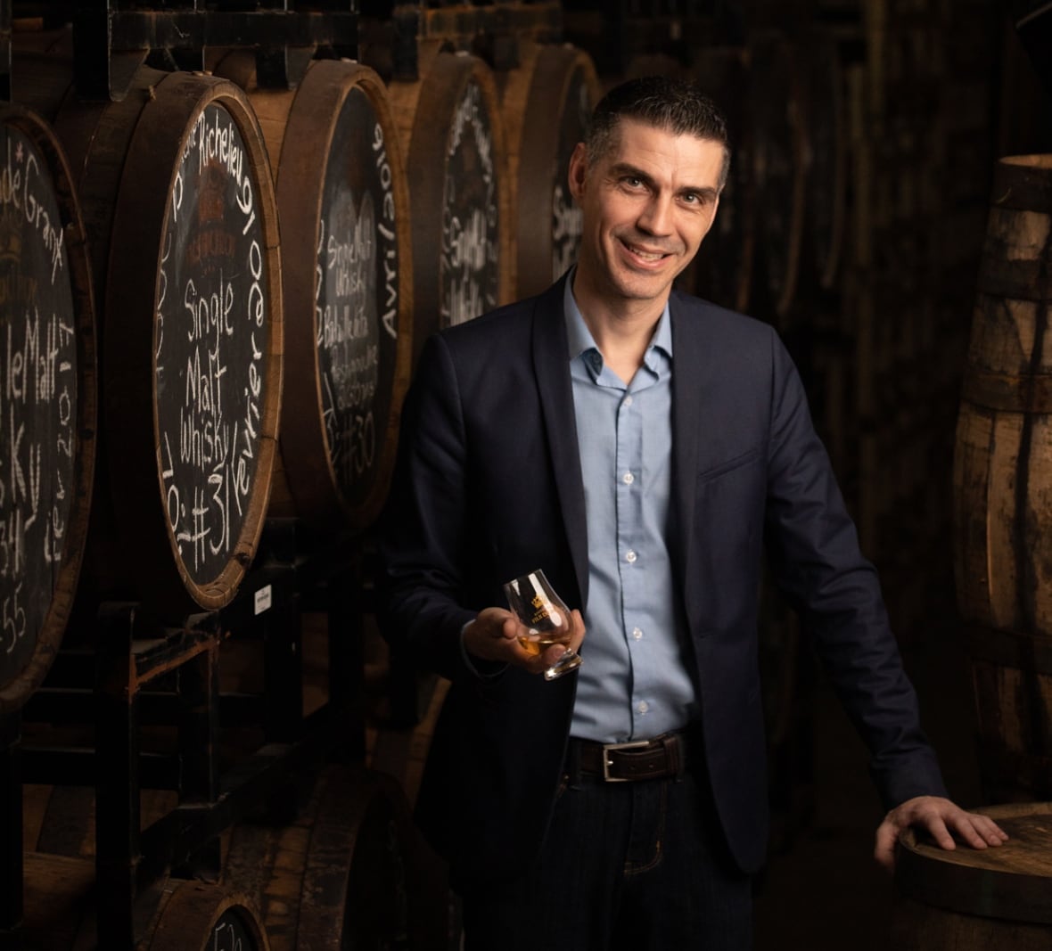 A man in a black blazer and blue collared shirt underneath holds a small glass in front of barrels at a distillery.
