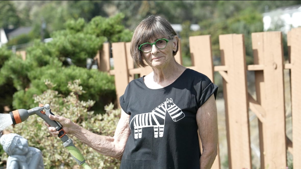 A woman with chin-length hair and green glasses holds a garden hose.