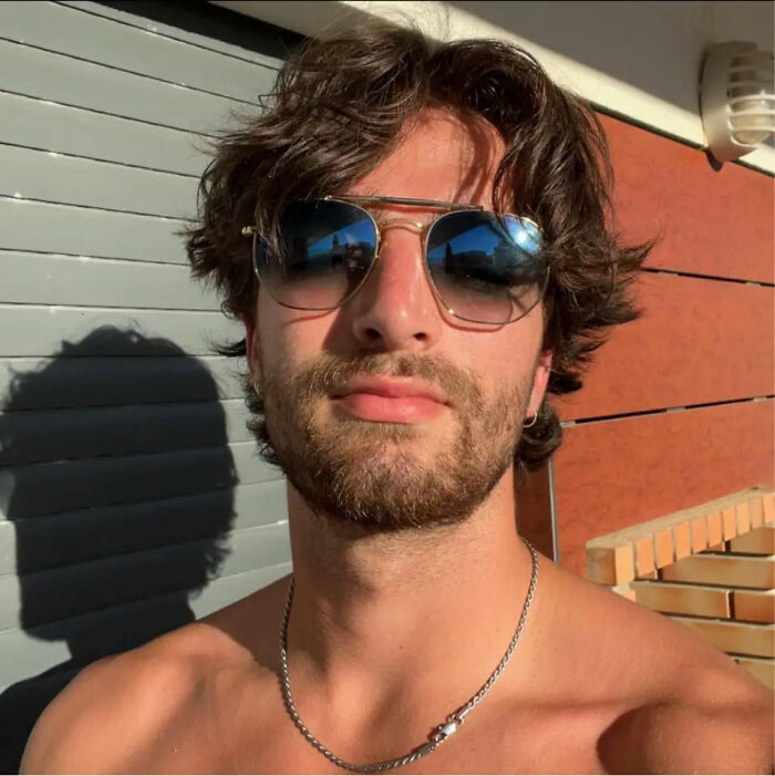 Young man wearing sunglasses and a chain necklace posing outdoors with sunlight and shadows in the background.