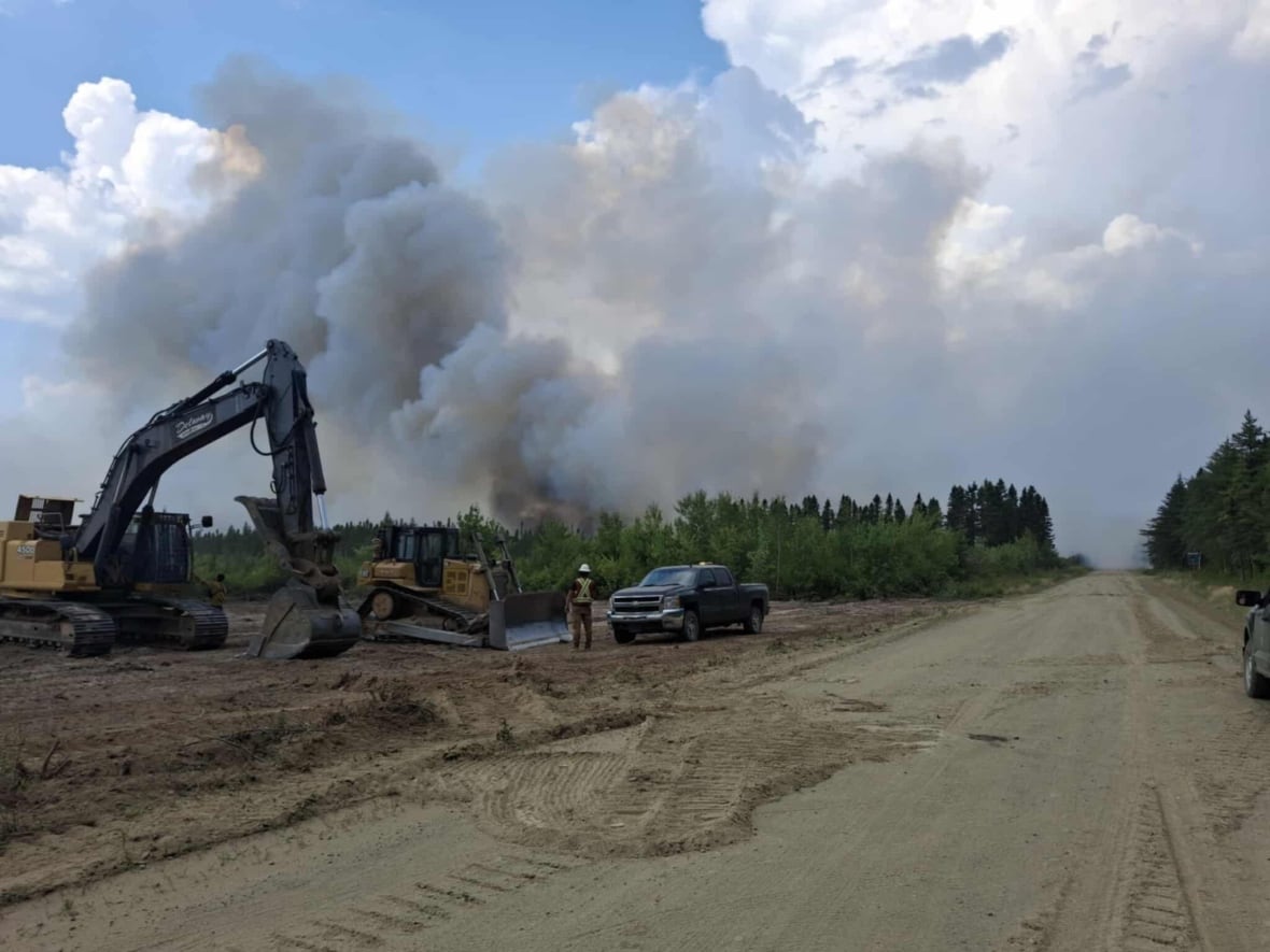 An excavator and small pick-up truck on a dirt road. In the distance, plumes of smoke fly into the sky.