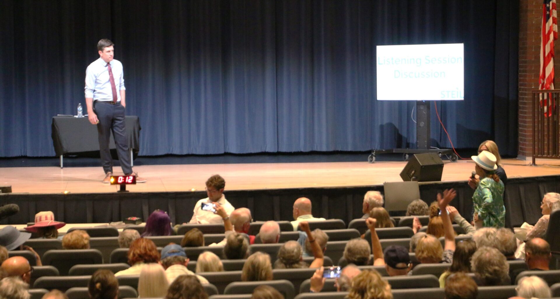 A man stands on stage during a listening session discussion, addressing an audience seated in an auditorium. A sign and an American flag are visible onstage.