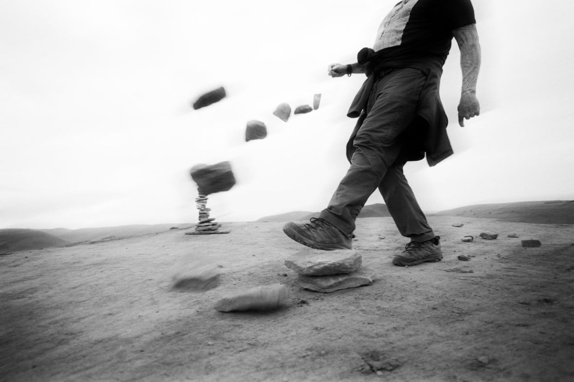 An artsy, blurry black and white photo of a man, shown from the shoulders down, kicking over a pile of stones.