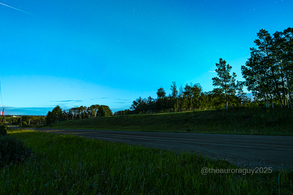 Meteors are visible streaking through a dark blue sky wreathed in a green aurora above a rural landscape.