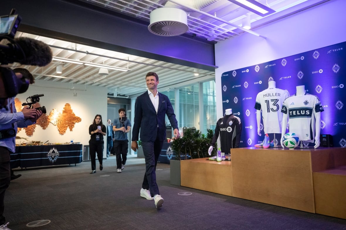 A sharp-dressed man walks in front of news cameras and team jerseys on display.