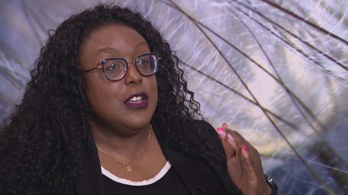 A middle aged black woman speaks to someone off screen. She is seated indoors, visible from the chest up, gesturing to the off-camera person.