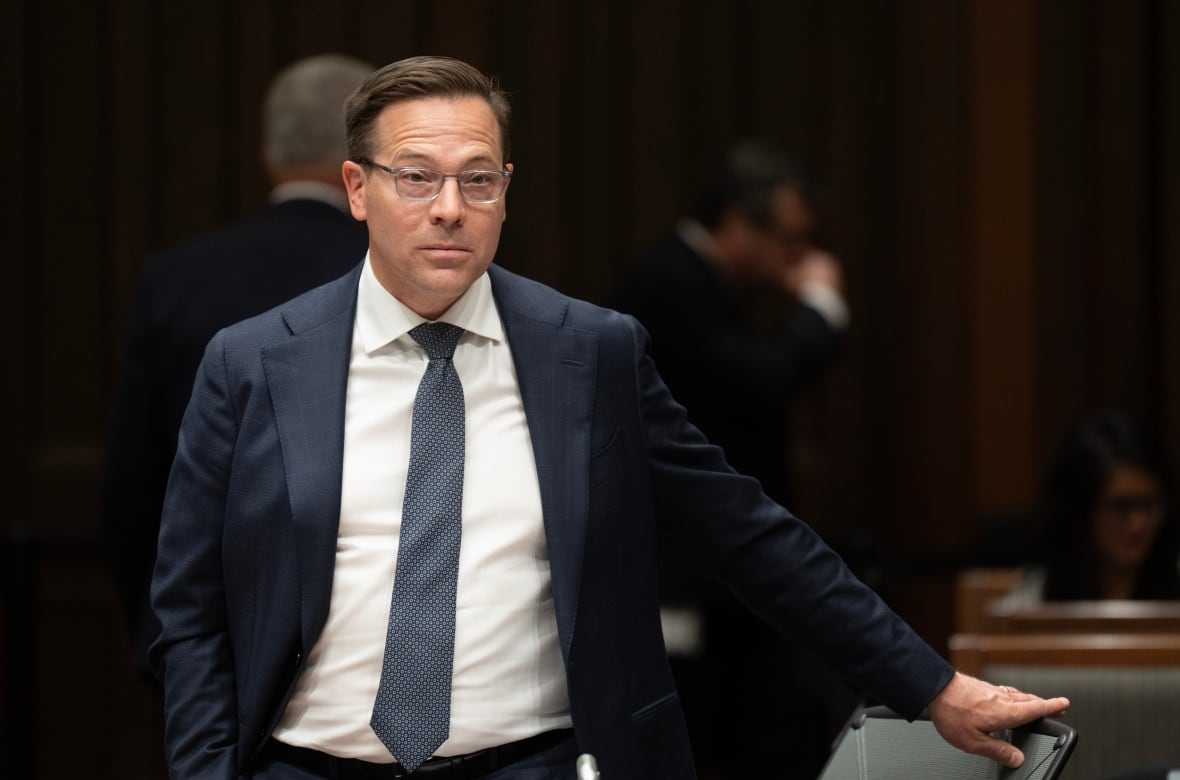 A man with glasses and wearing a suit stands in a meeting room with his left hand on the back of a chair.