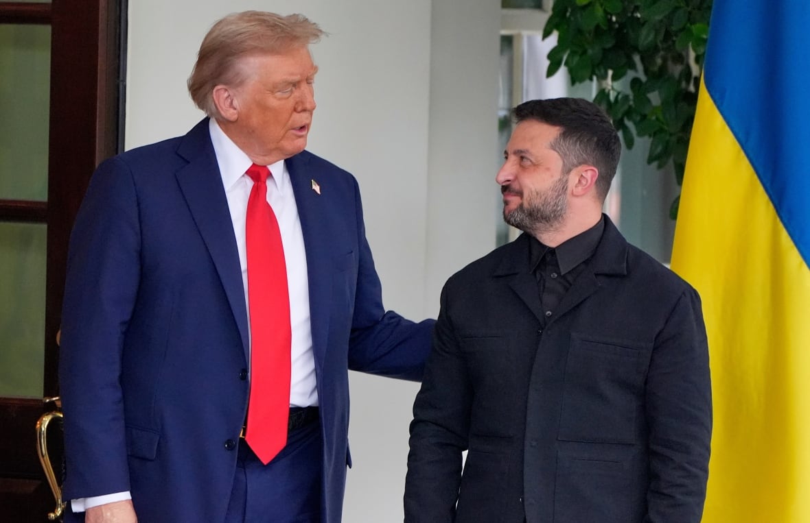 A man wearing a blue suit and a red tie touches the arm of a bearded man wearing a black suit.