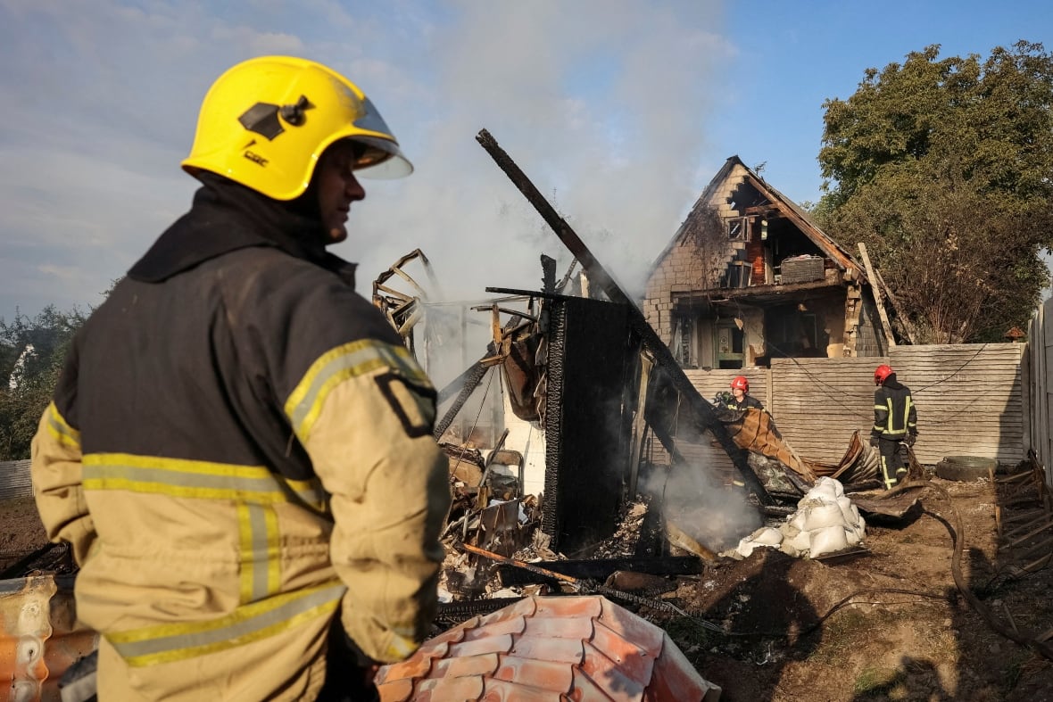 A helmeted firefighter is shown in profile in the foreground as colleagues work at the site of a damaged and charred residential building.