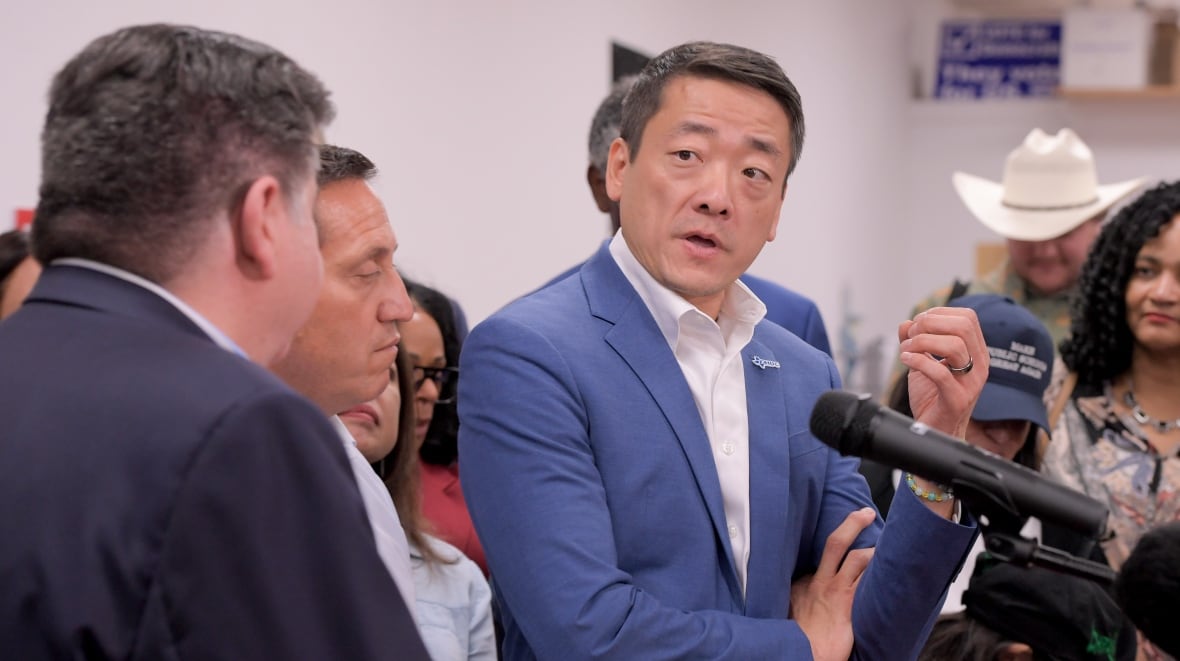 A man in a blue suit stands in front of a microphone, but looks off to the side to talk to another man standing near him. 