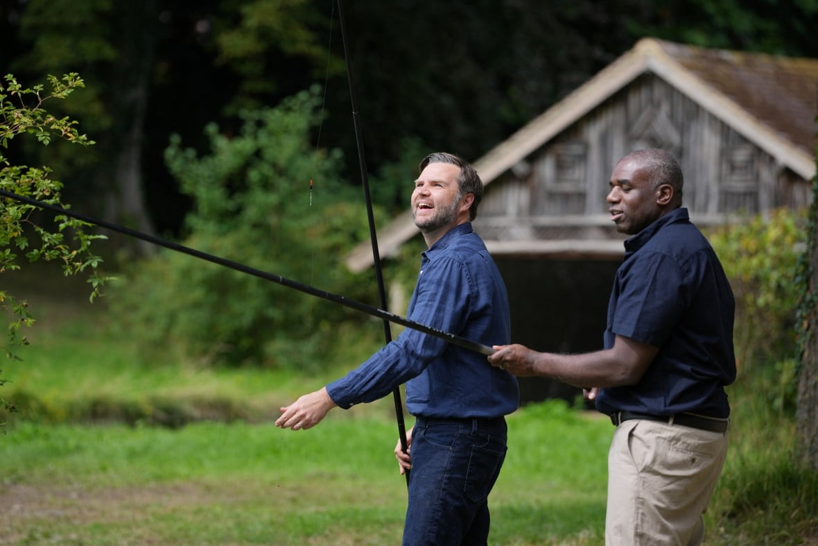 A smiling, bearded Caucasian man and a cleanshaven dark-complected man fish in a bucolic setting.
