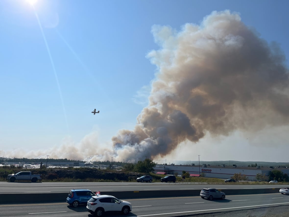 A plane is seen in the sky next to a large plume of smoke over a wooded area. 