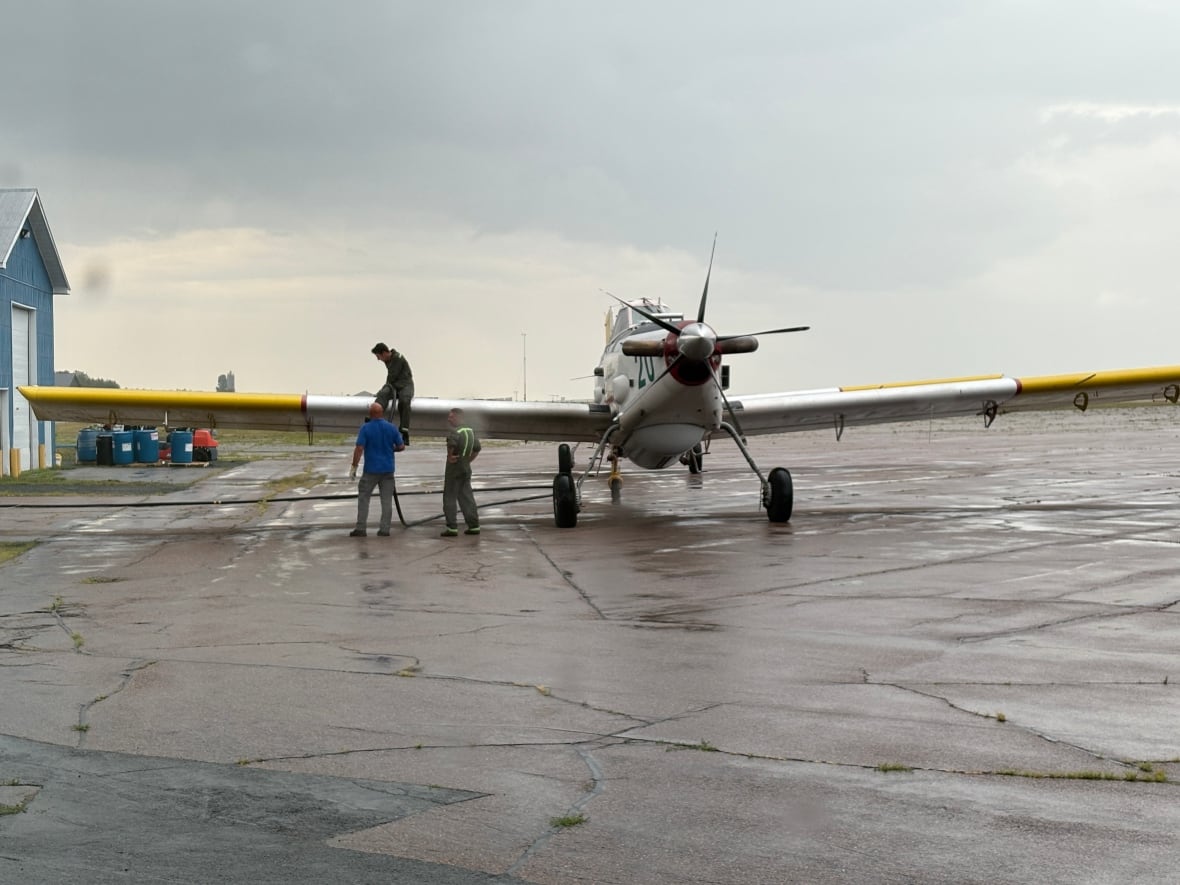 A man sitting on a plane wing as other men help refuel it. They're all on a tarmac in the rain. 
