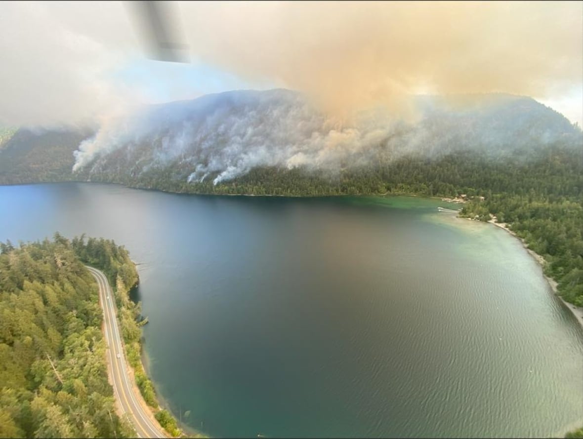 Orange smoke arises from a forested hill near a picturesque lake.