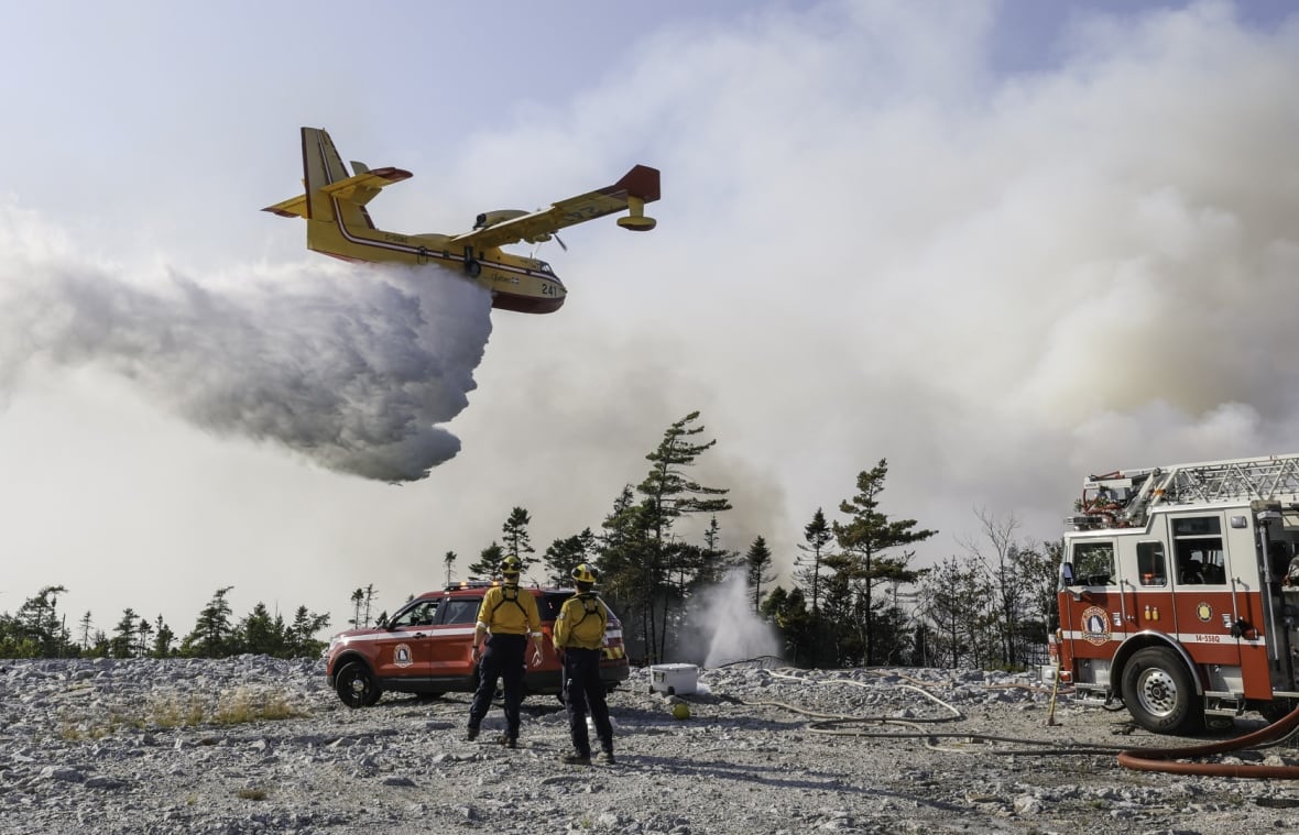 A water bromber drops water over a wildfire as two men in fire fighting gear look on.