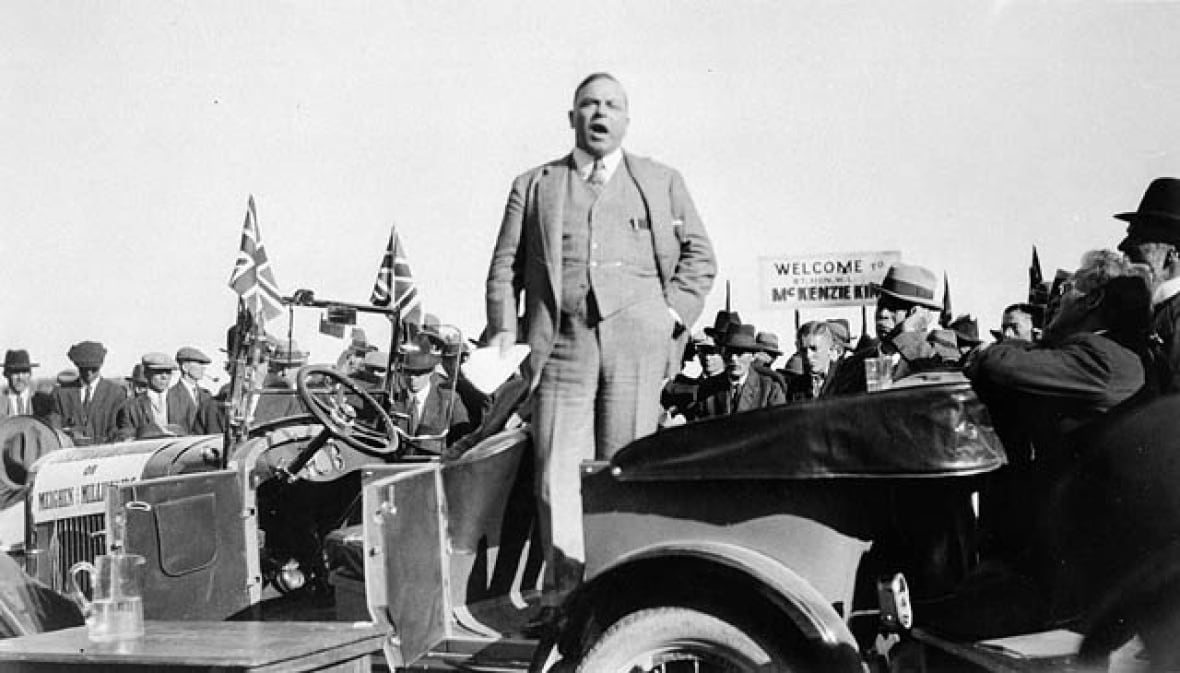 An old black and white photo shows a man in a suit standing in a 1920s style care speaking to a crowd.