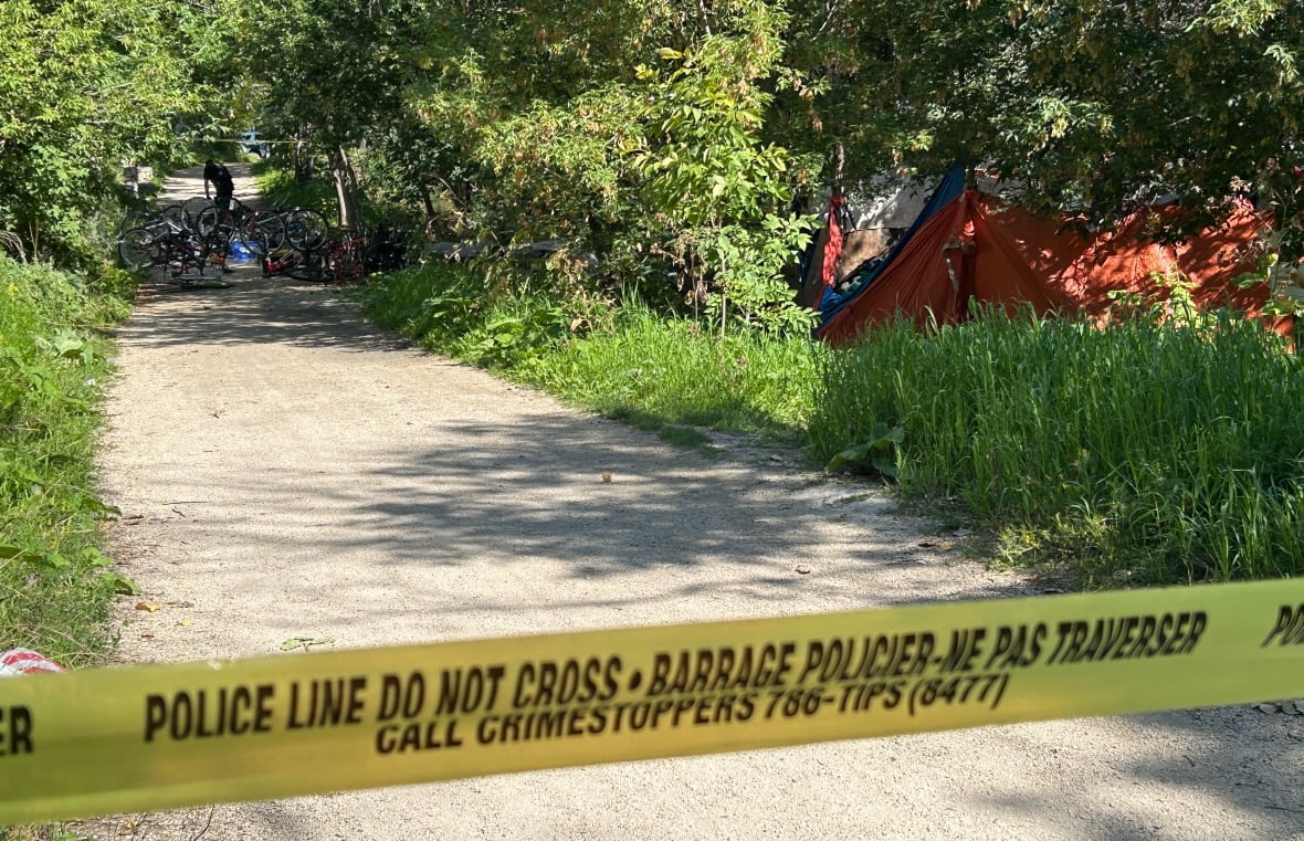 Police tape is seen across a gravel bike path, with a tent and several bicycles on the path in the backgroun.