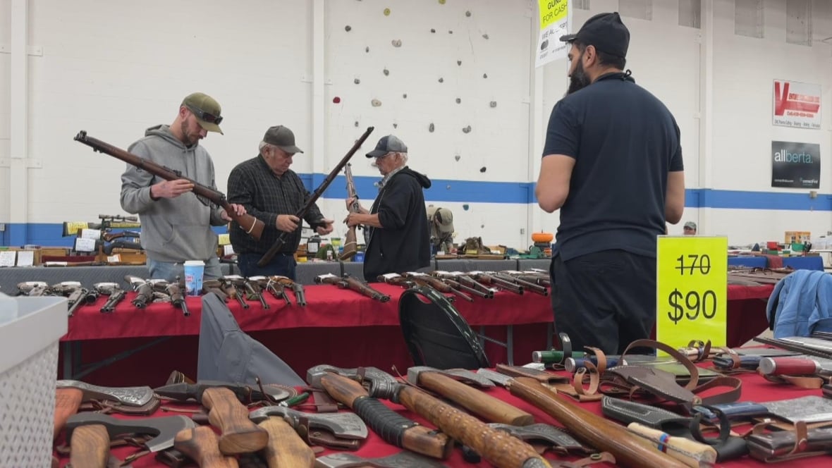 A man stands in front of tables where people are looking at guns.