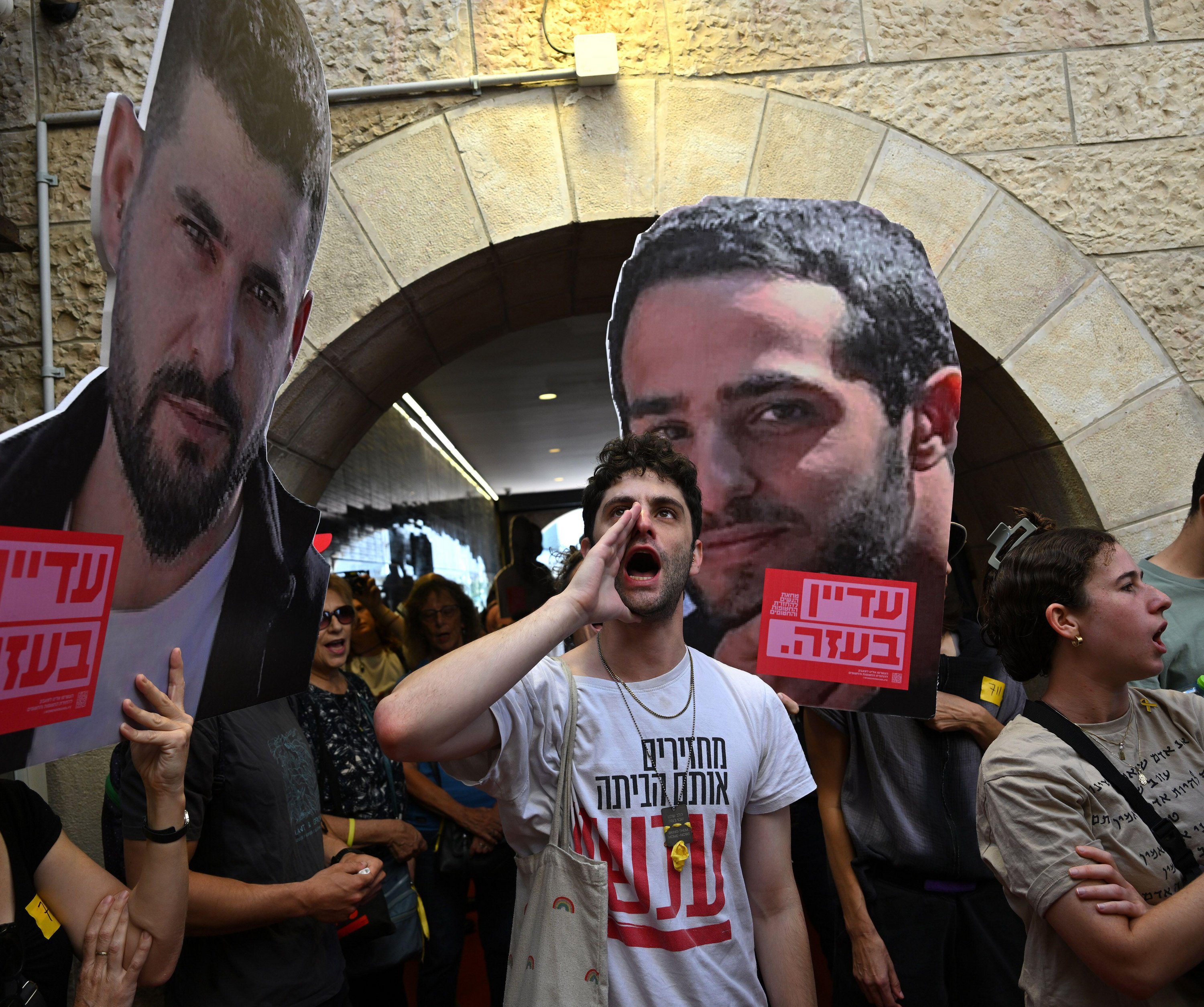 A man shouts at a protest in Jerusalem, holding large cutouts of hostages' faces, demanding the end of the Israel-Hamas war and the return of all hostages.