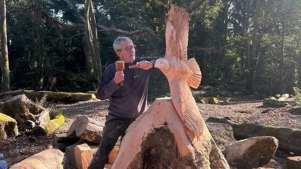 A man wearing dark clothing uses a chisel to carve a large bird into a tree trunk in a forest