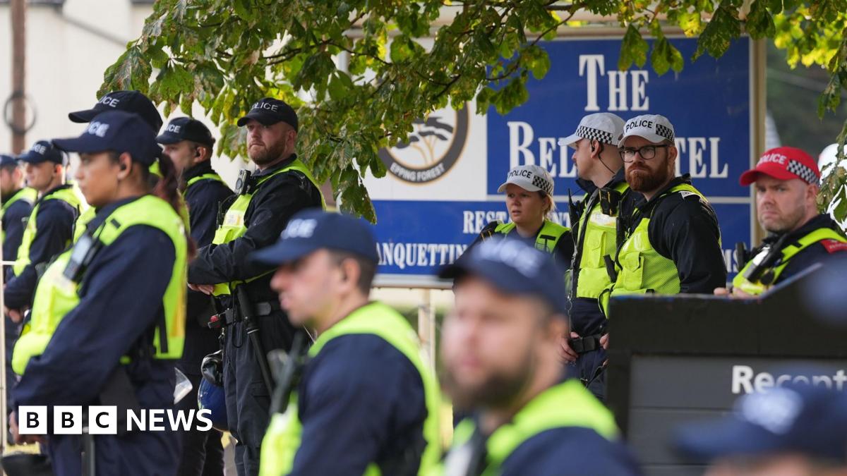A large group of police officers, all wearing caps and hi-vis vests, standing in front of a large blue sign outside The Bell Hotel, which advertises its name.