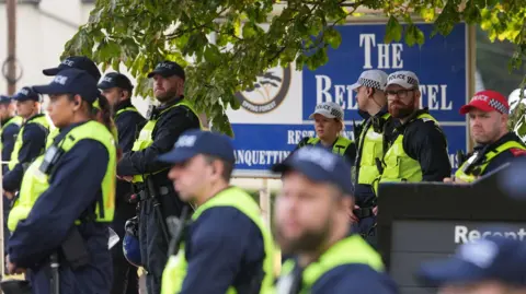PA Media A large group of police officers, all wearing caps and hi-vis vests, standing in front of a large blue sign outside The Bell Hotel, which advertises its name.