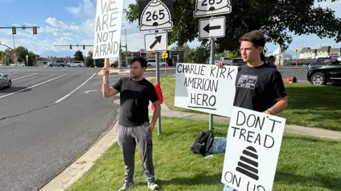 BBC / Christal Hayes McKinley Shinkle (left) and his cousin, Anthony, hold signs in support of Charlie Kirk near the entrance of the Utah Valley campus 