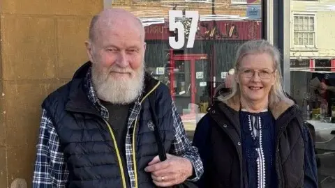 Family handout Peter on the right of this image is shown after a recent hair cut back in the UK. His beard is trimmed and his hair shorter. His wife, Barbie, stands smiling next to him.