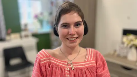 A head and shoulders shot of Lily. She has a pink and white top on, and a silver pendant necklace. She smiles at the camera and has short brown hair. 