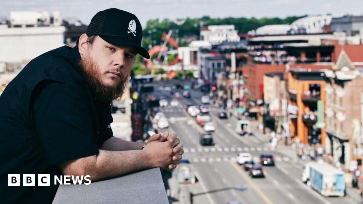 In this image released on June 10th, 2021, Luke Combs poses on set for the 2021 CMT Music Awards in Nashville, Tennessee. He is dressed in black with a baseball cap and is overlooking a busy street.