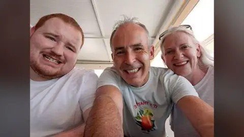 Family photo Evan O'Connor and his parents Mark and Louise O'Connor smiling at the camera in a family photo.   Evan has short, ginger hair and a beard.  Mark and Louise both have grey hair.  They are all wearing white t-shirts. 
