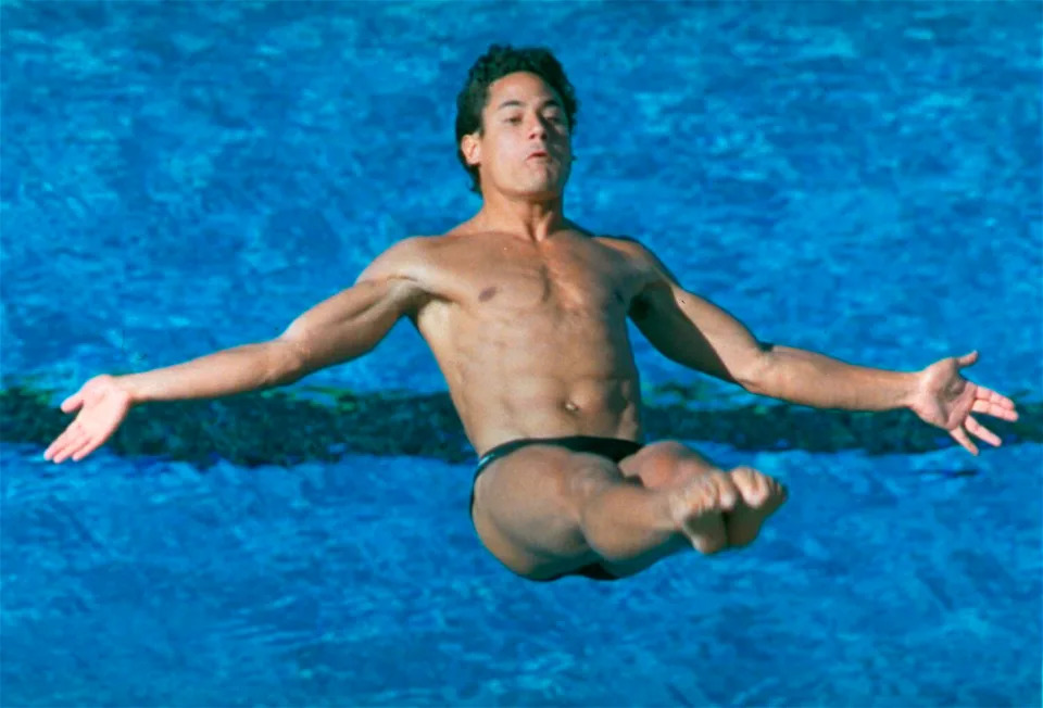 Greg Louganis spreads his arms and bends at the waist while in mid-dive over the water