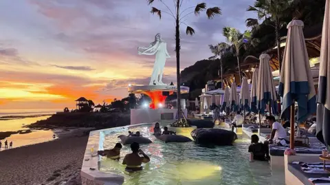 Getty Images Sunset in the background of a beach, as people relax in a pool at a beach club. There are sunbeds and floaties at the beach club.