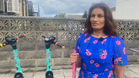 BBC Anela Wood, a blind woman, is wearing a royal blue dress with pink flowers and holding a pink walking cane. There are two turquoise e-scooters parked on the pavement next to her. She has dark brown hair and is wearing a gold necklace.