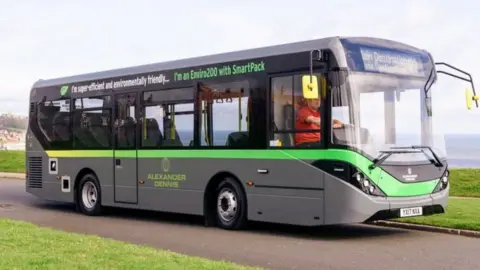 Alexander Dennis a grey electric bus with a green line and writing across it. It has an Alexander Dennis logo on the side.