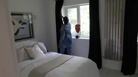 Patricia Ogunfeibo in one of the bedrooms of the flat she owns, drawing a curtain. There is a double bed with light coloured bedsheets, a large painting over the bed, a mask shaped ornament on the windowsill, the curtains are a dark blue/black and there is a chrome upright radiator to the right of the window.
