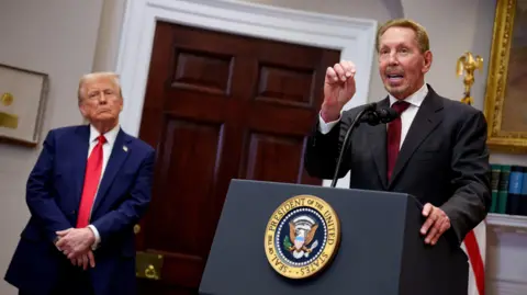 Getty Images Larry Ellison at the White House with Donald Trump in January 2025. Ellison is at a podium in a dark suit with burgundy tie with one hand up for emphasis while Trump stands on the side, with his hands folded in front of him and his head cocked