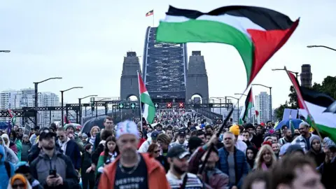 Getty Images A large crowd of people, some waving Palestinian flags, walk toward the camera with the Sydney Harbour Bridge behind them