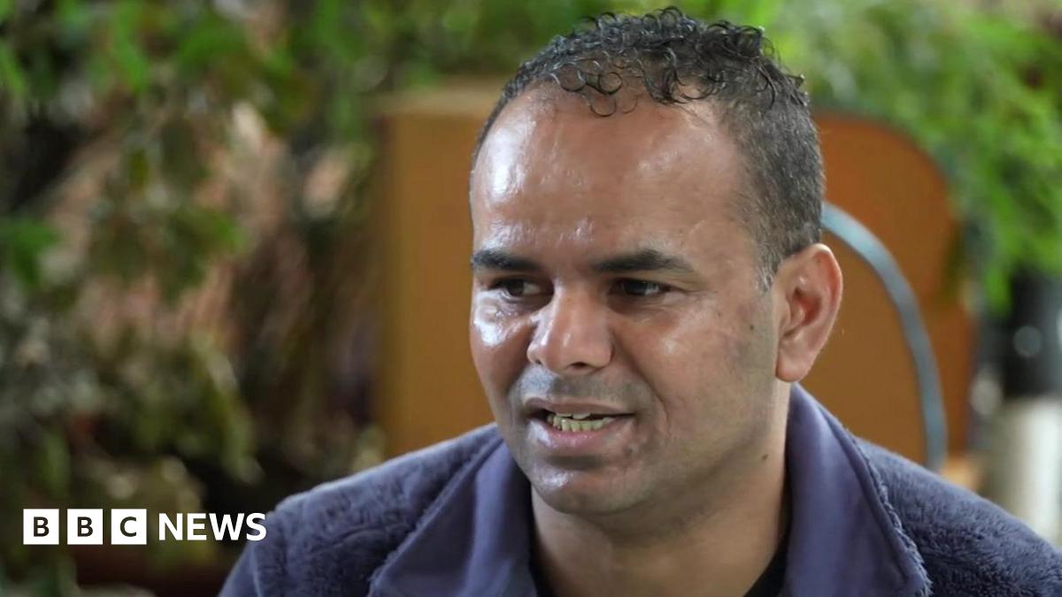 Mohamed has short black curly hair and stubble. He is wearing a blue jumper and is speaking to a BBC reporter interviewing him, who is out of shot.