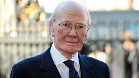 Getty Images Sir Menzies Campbell who has greay hair and glasses. He is wearing a dark suit and tie with a white shirt. He is standing in front of a blurred Houses of Parliament