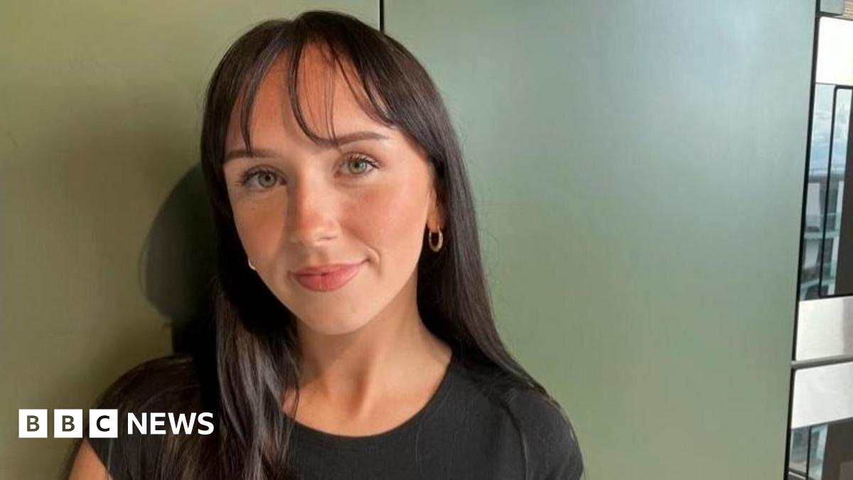 Leoni Clancey, 21, with shoulder length straight dark brown hair with a wispy fringe wearing a black t shirt standing against a khaki wall with a neutral expression
