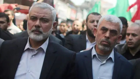 Getty Images Ismail Haniyeh (left) and Yahya Sinwar (right) wear open-necked shirts with a dark suit jacket amongst a crowd of people on a busy street. 