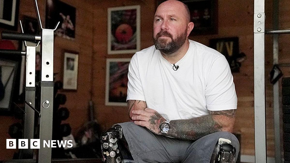 Dave Watson is sitting on a bench in his gym at home. He is wearing a white t-shirt and grey shorts. He has two dark black prosthetic legs covered in stickers. Behind him on the wall are photographs including one of him winning shootout gold medal at the Invictus Games in Sydney.