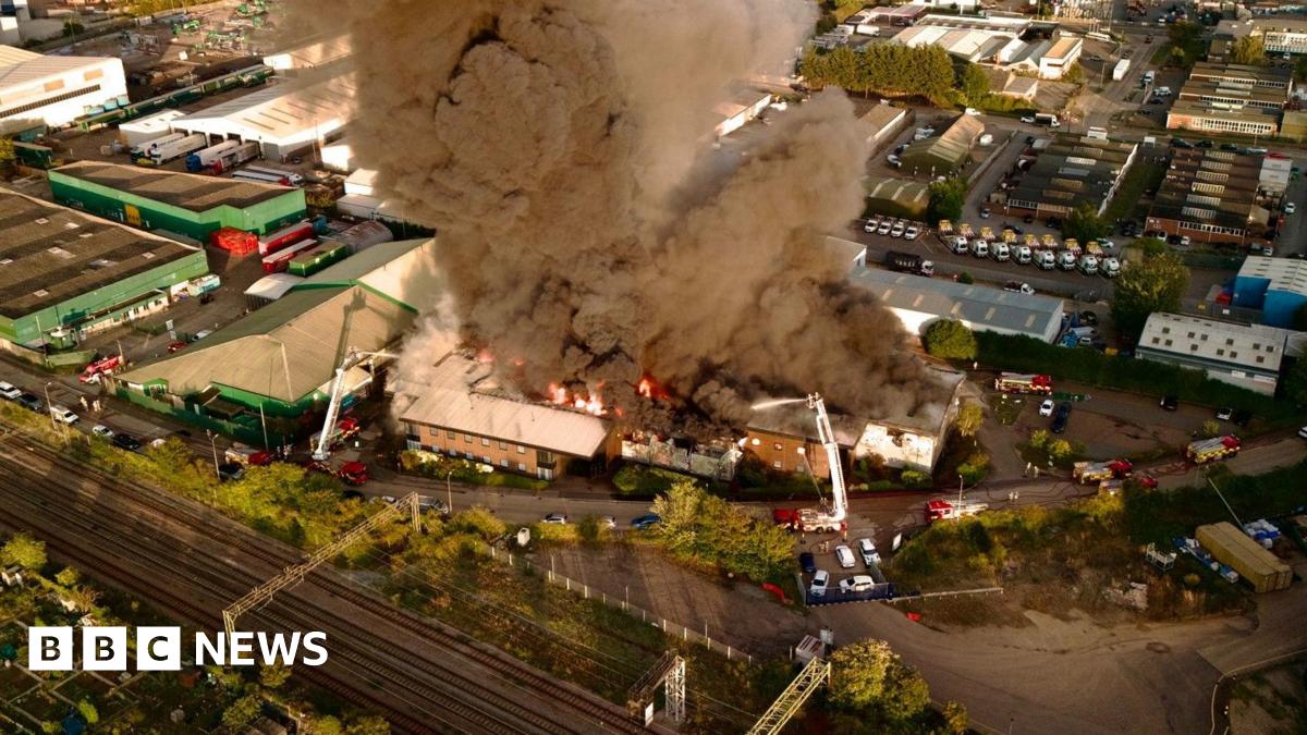 An aerial drone photograph above an industrial fire in Witham. Black smoke is billowing into the air and orange flames are coming off the roof. There are numerous fire engines at the scene with aerial platforms.