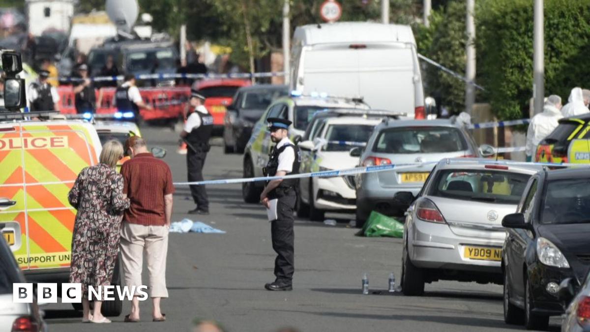 Members of the public look on from in front of blue and white police tape as police officers stand in a road filled with police vans and cars, with debris strewn across it.