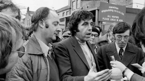 Getty Images A black and white shot of a man speaking to journalists - who are out of the shot. He is surrounded by other men and is standing in front of a sign which reads "Marathon shipbuilding co"