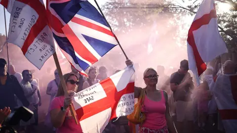 AFP via Getty Images A group of people gathered outdoors in a wooded area, holding flags and signs. Visible flags include the Union Jack and the flag of England. Signs read 'Protect Our Children' and 'Keep Our Children Safe.' Sunlight filters through the trees, creating a hazy, backlit atmosphere.