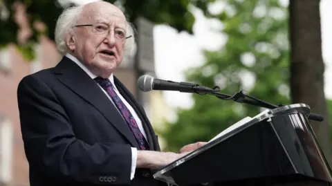 PA Media Michael D Higgins standing at a lectern he is wearing a black suit with a black tie that has purple flower patterns on it. He has glasses on. 