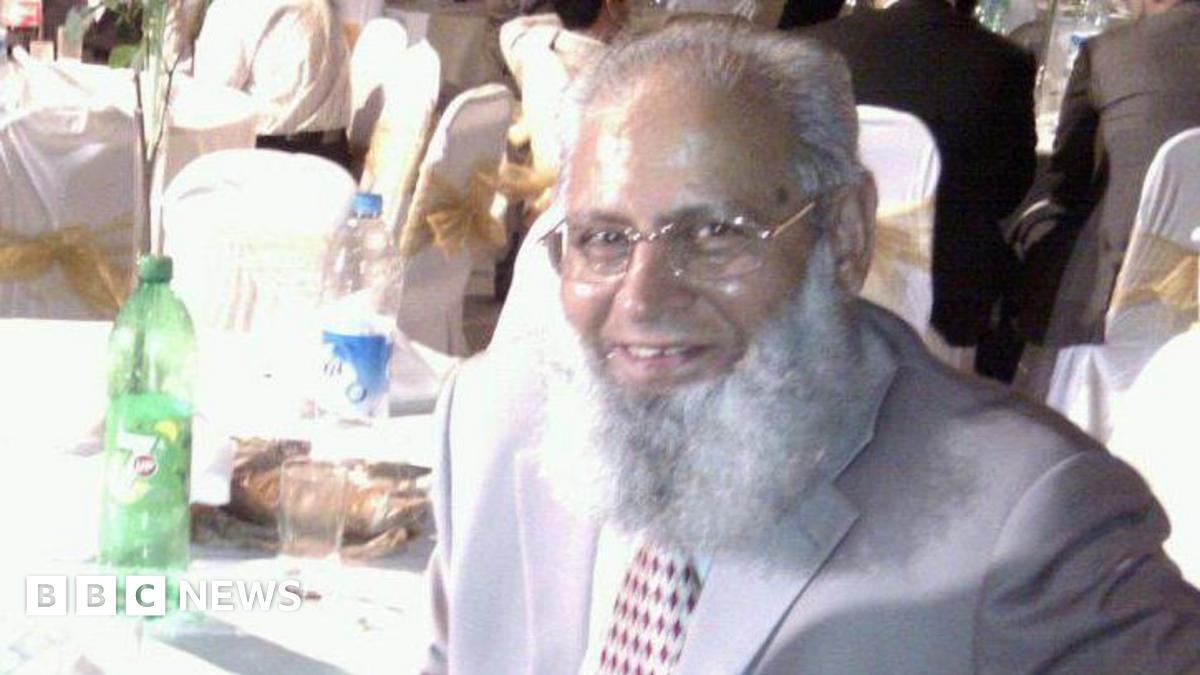 A man in thin framed glasses with a white beard smiling as he sits at a table during a celebration. He is wearing a grey suit and red and white patterned tie, and drinks can be seen on the table behind him.